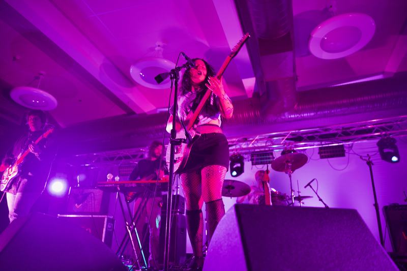 caption: Karly Hartzman, as Wednesday’s frontwoman, sings into the standing mic as she looks down at the guitar she is playing. Photo taken by Molly Shoffner for Wavelength.