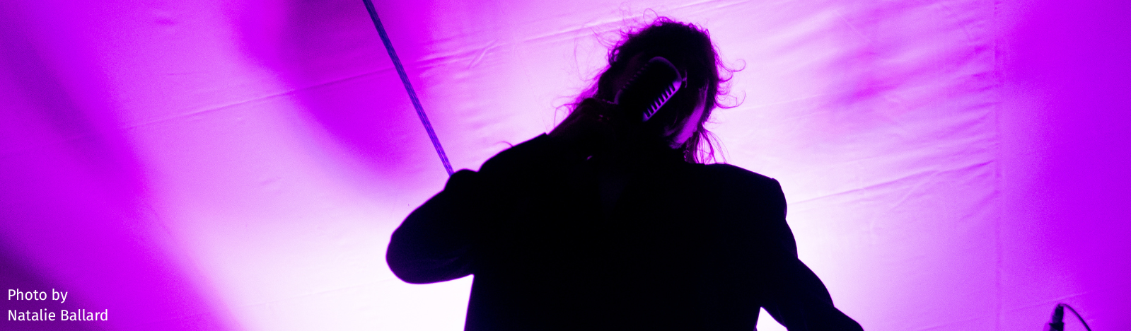 A photo of a singer in silhouette during a concert with a background that is lit up with purple light from the stage lighting. The photo is of the band Thunderpussy and was taken by photographer Natalie Ballard.