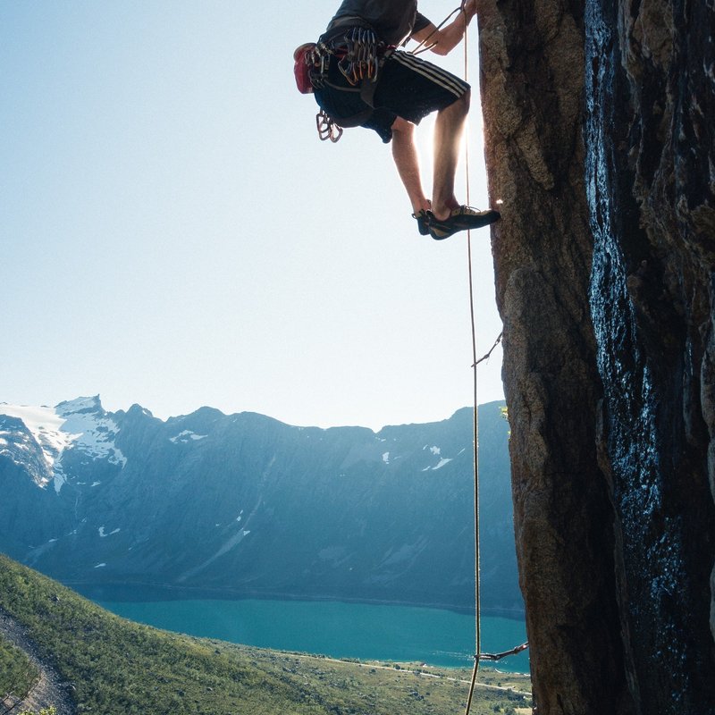 Student climbing cliff face with mountains, forest and lake in the background.