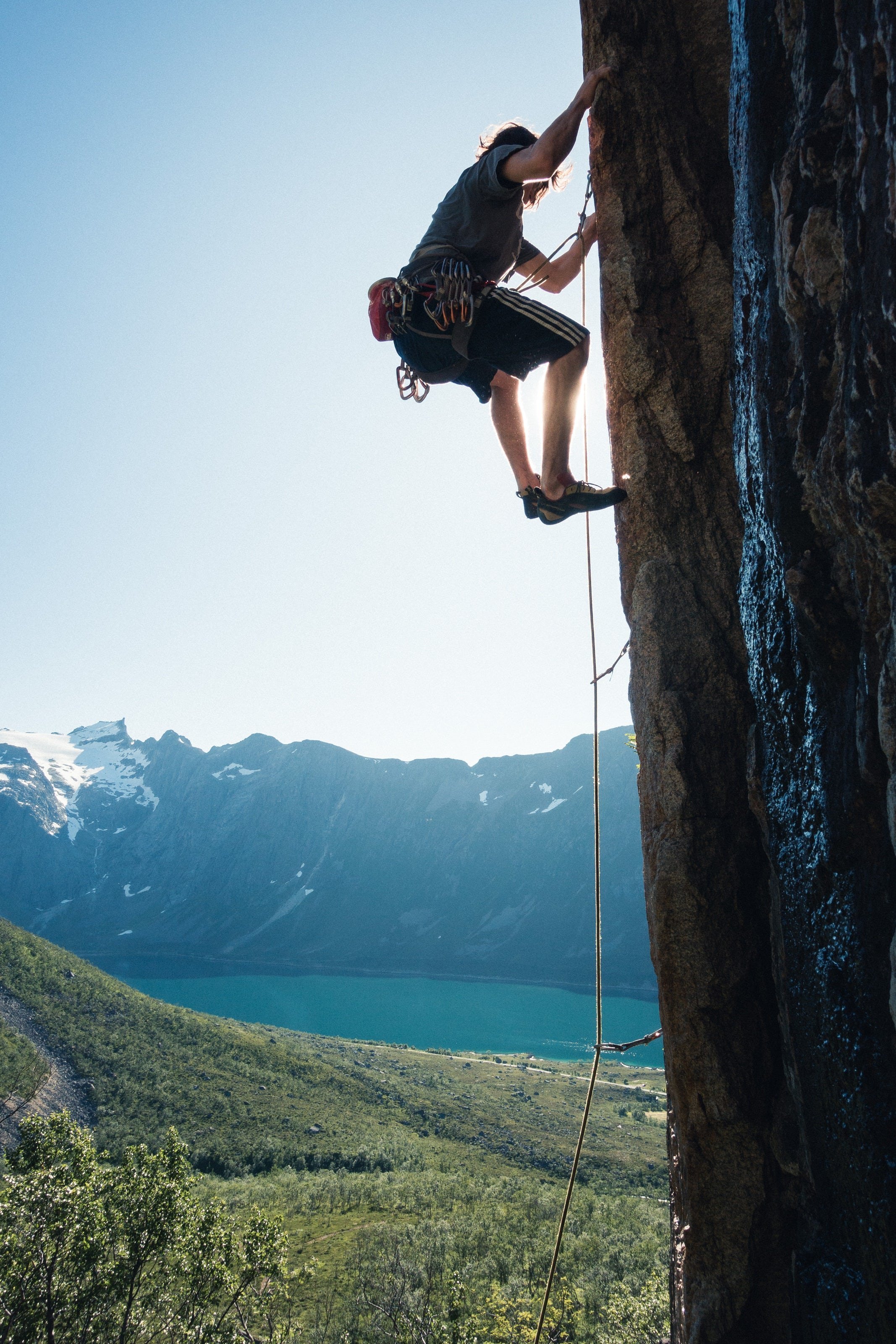 Student climbing cliff face with mountains, forest and lake in the background.