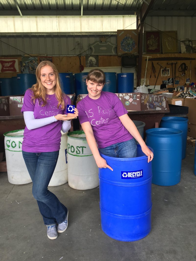 Students with A S Recycle Center shirts hold a recycle sticker and stand in a large blue recycle bin