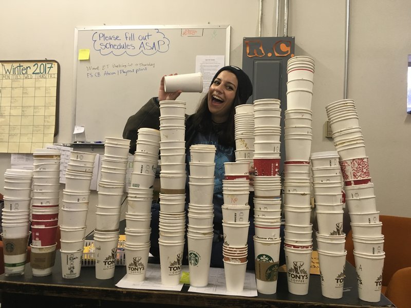 Student pretends to drink from a paper coffee cup. Many stacks of similar cups are in front of them