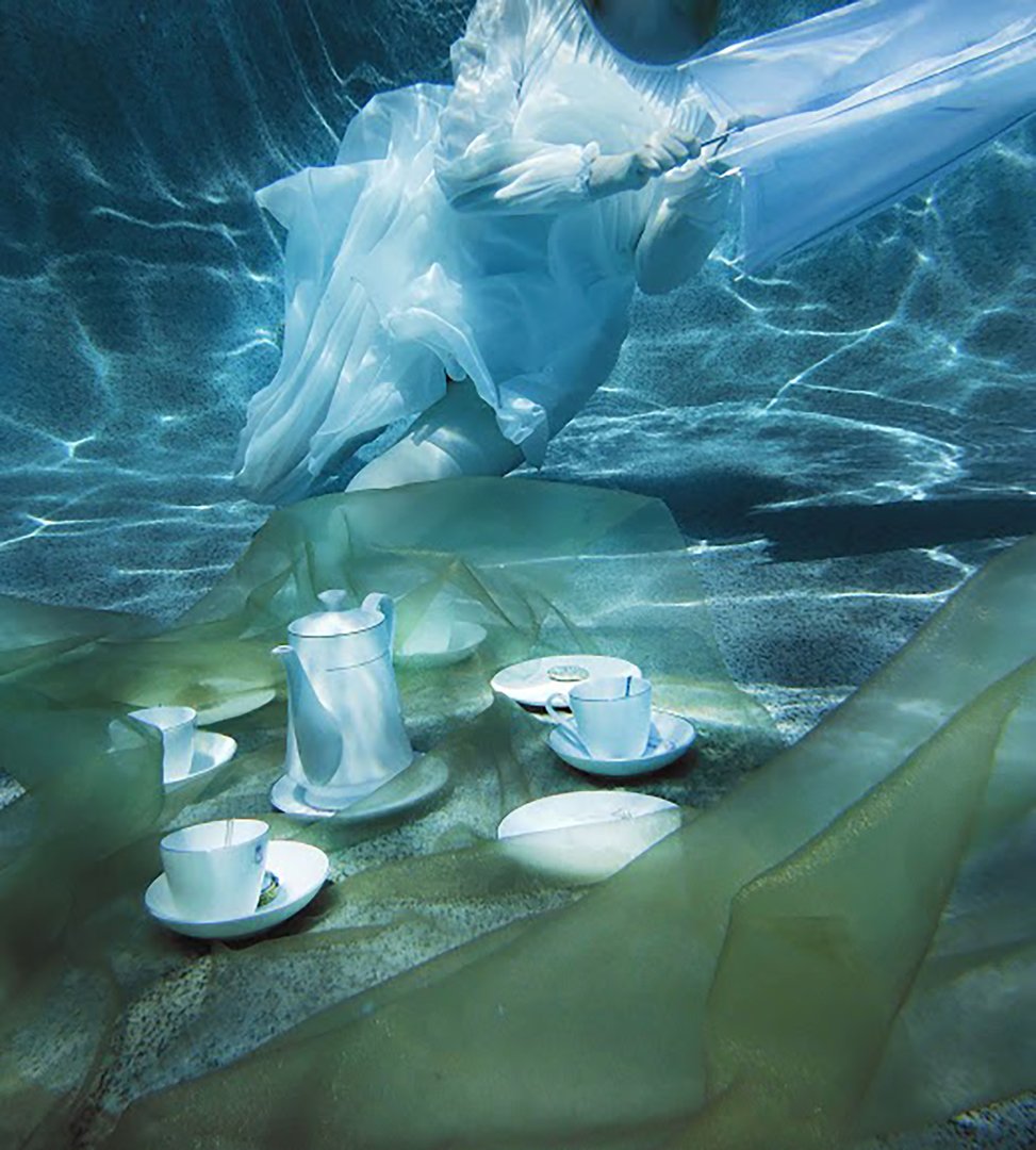 Underwater photo of a figure in a white dress with an umbrella sat by a tea set