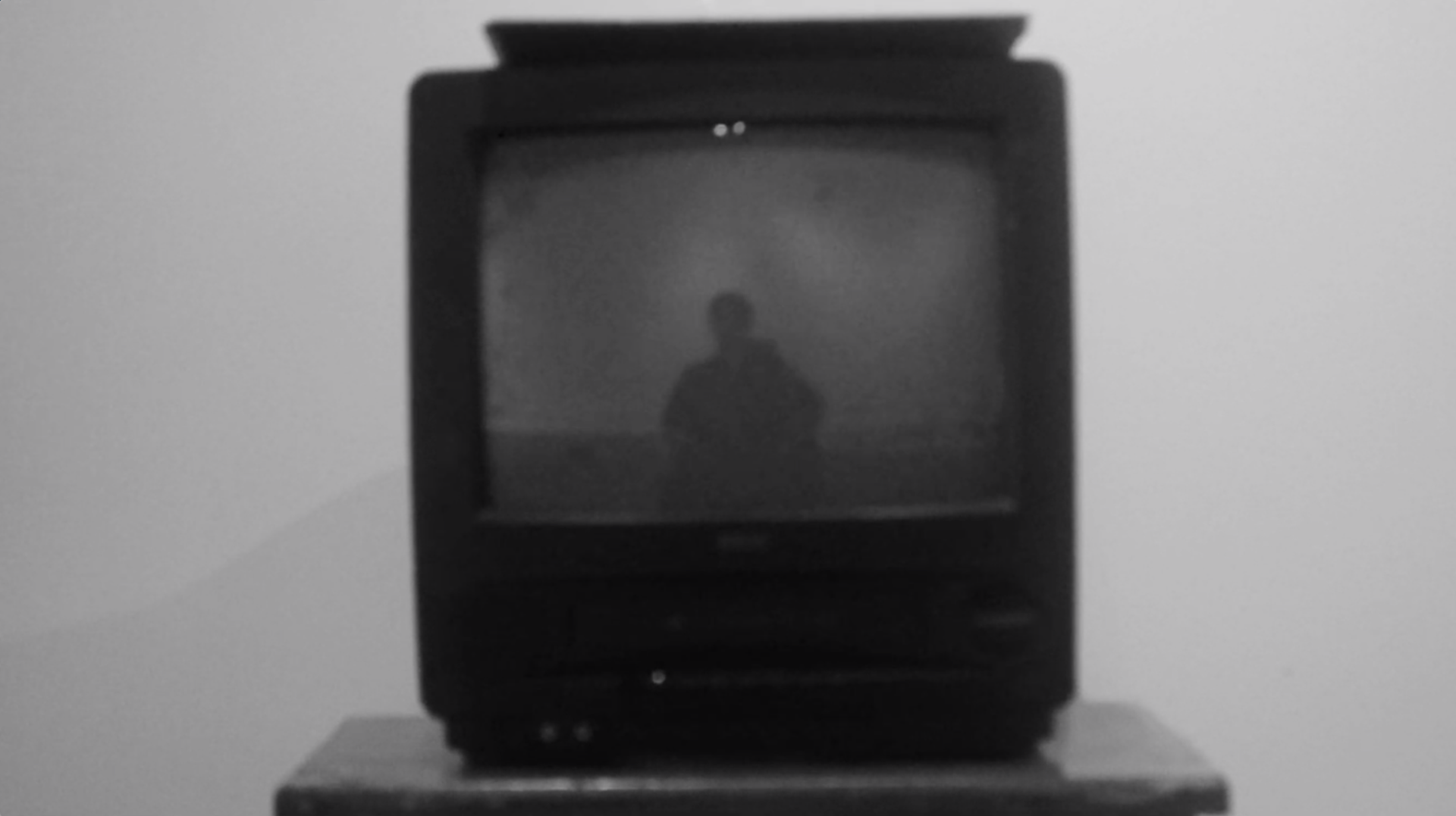 Screen capture of a digital short film which shows a black and white scene of a box television reflecting the actor who is sitting in a chair
