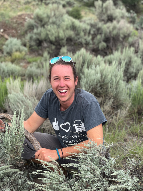 Ben Crandall smiling at the camera in a field of sage brush wearing teal sunglasses on top of his head