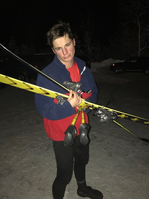 Grant Rienstra looks disapointedly at the camera as he holds his skiis and poles in a messy bunch that will surely fall appart soon. He is in a dark and snowy parkinglot with crocs on